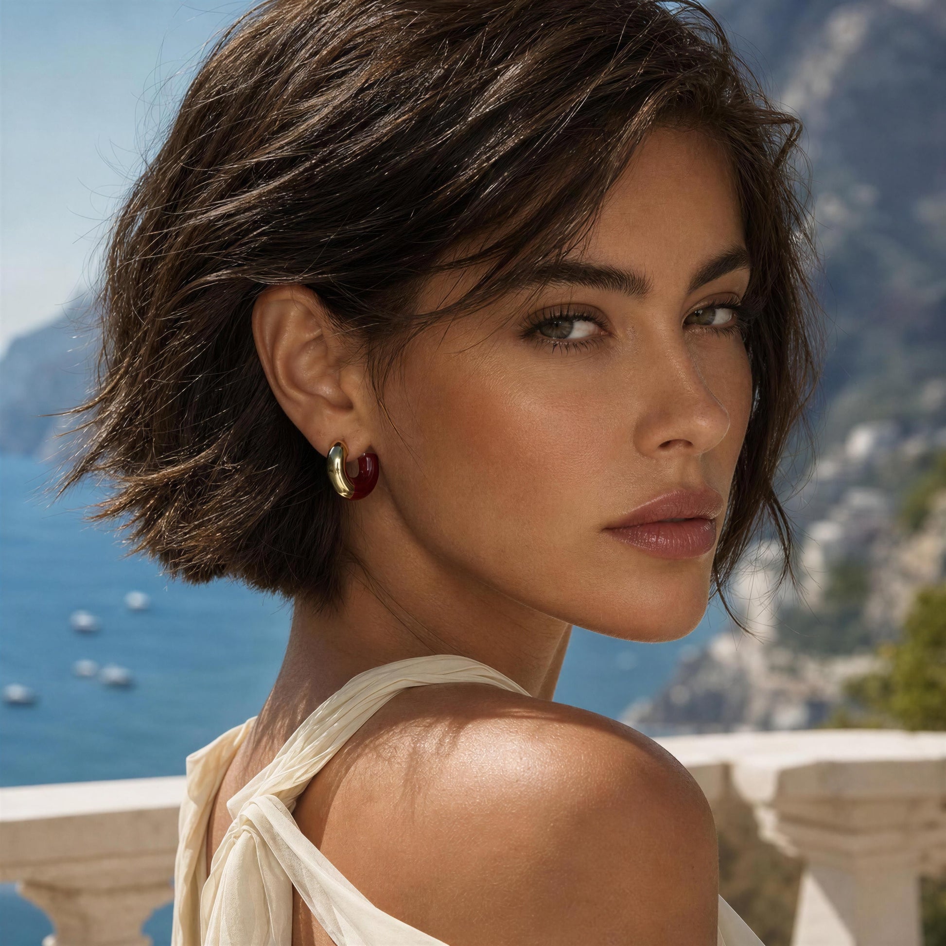 A woman with a short dark brown bob hairstyle wearing a gold and burgundy crescent hoop earring, the Gatsby Arc Earrings from Chica Chica Jewelry, photographed outdoors with a scenic blue sea and mountains in the background.