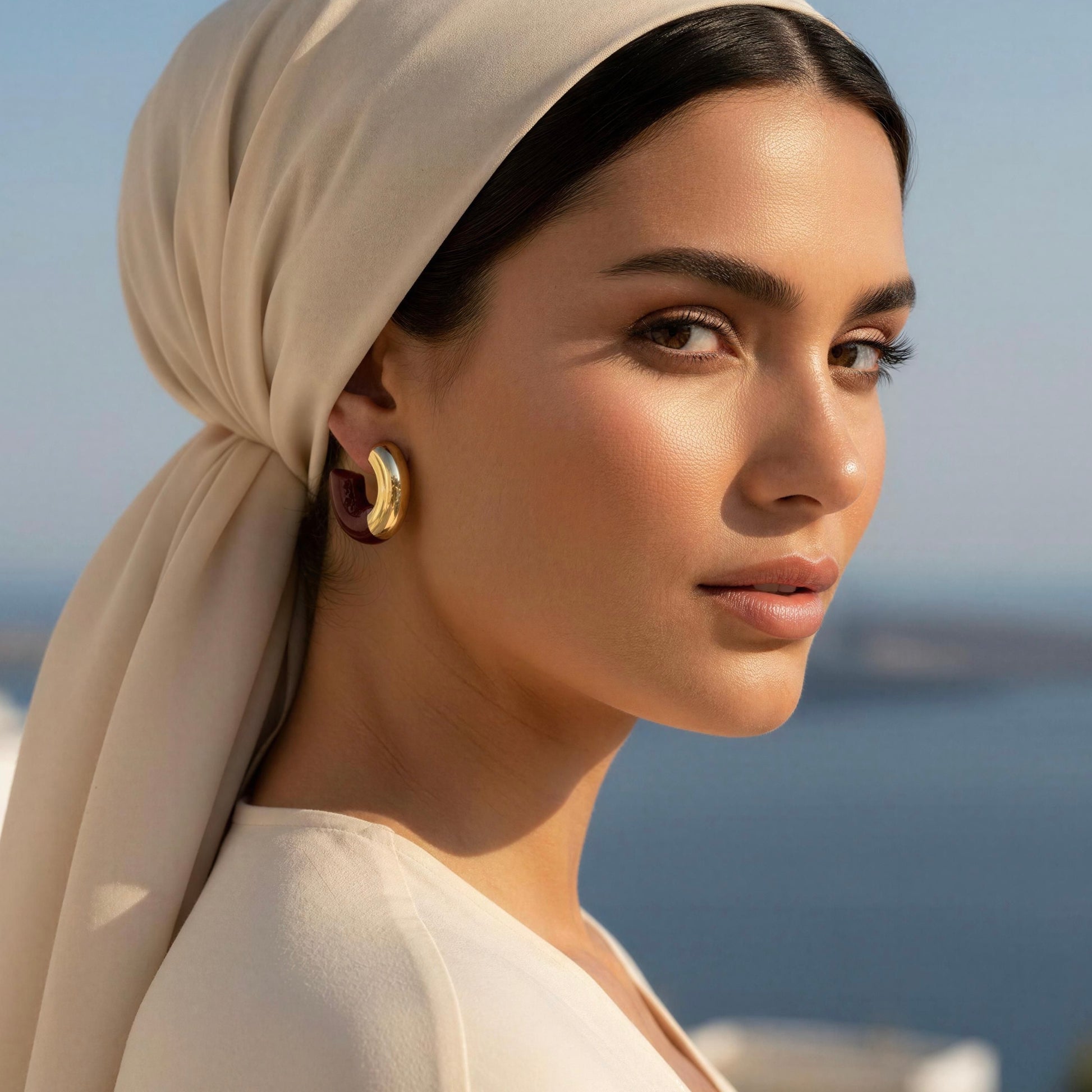A woman in a beige headscarf and cream top wearing the Gatsby Arc Earrings in Merlot color from Chica Chica Jewelry, set by Mediterranean terrace.