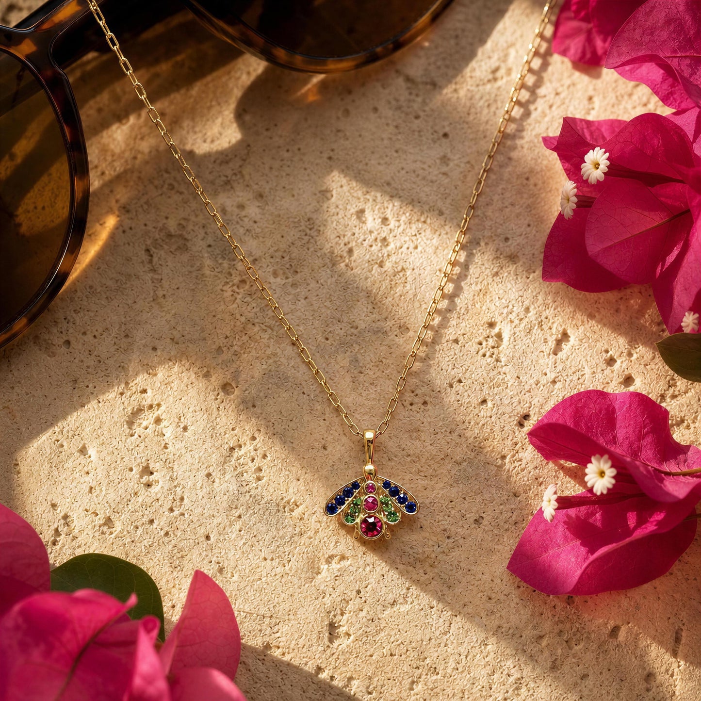 The Golden Meadow Bee Necklace, styled on a textured beige stone surfacpink flowers and sunglasses in the background.