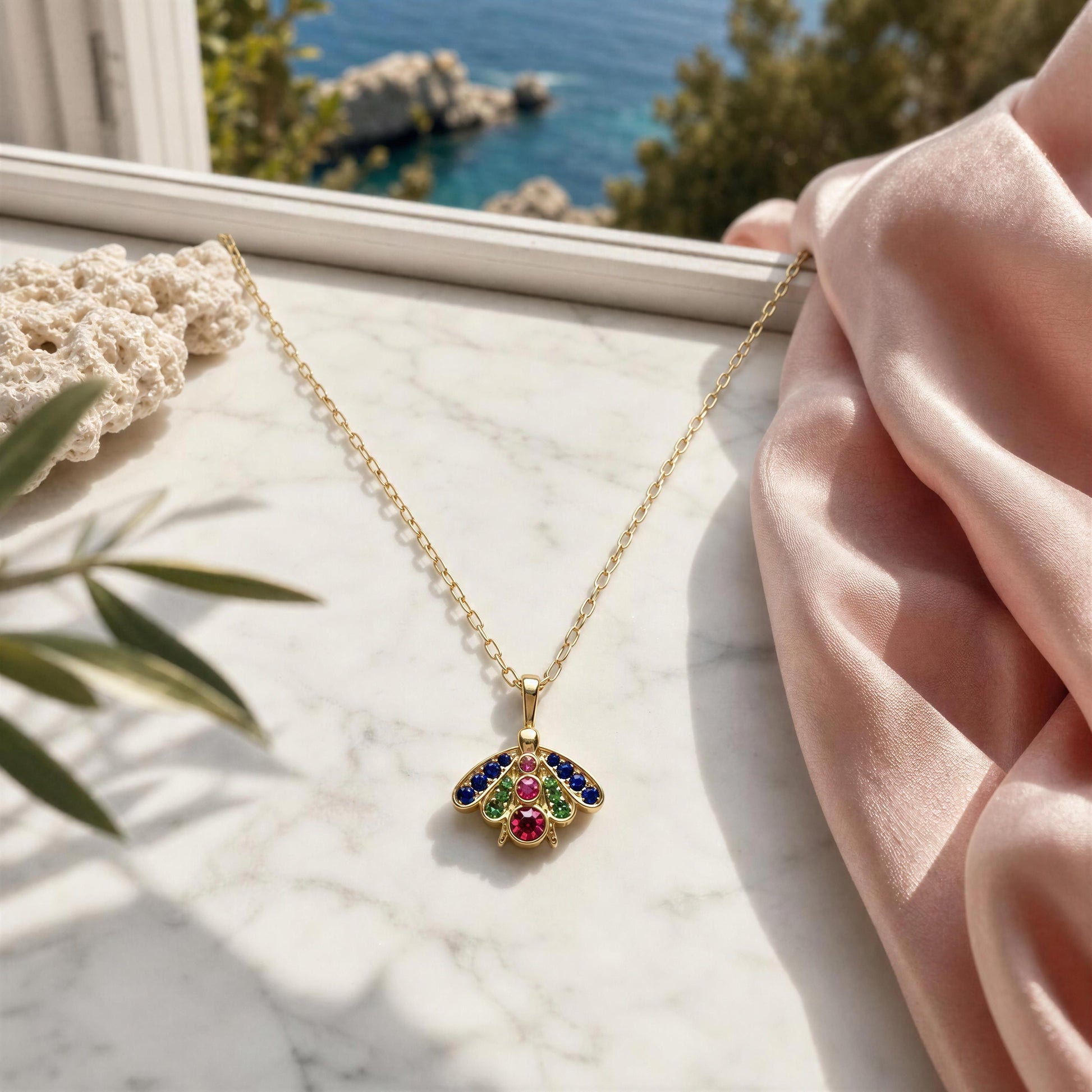 A gold chain necklace with a butterfly pendant, decorated with blue, green, and red gemstones in the center, displayed on a white marble surface with a pink satin cloth and a coastal background.