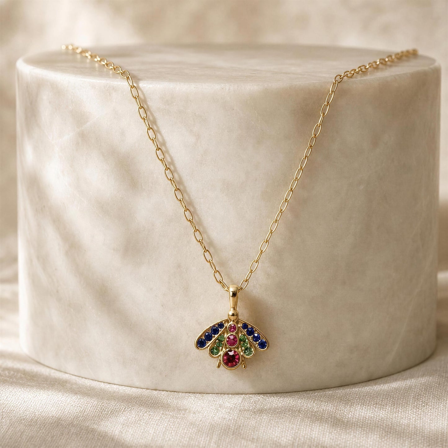 A gold chain necklace with a butterfly pendant, displayed on a textured white cylindrical pedestal. The butterfly's wings are embellished with blue, green, and red gemstones, and its body is accented with a central red stone.