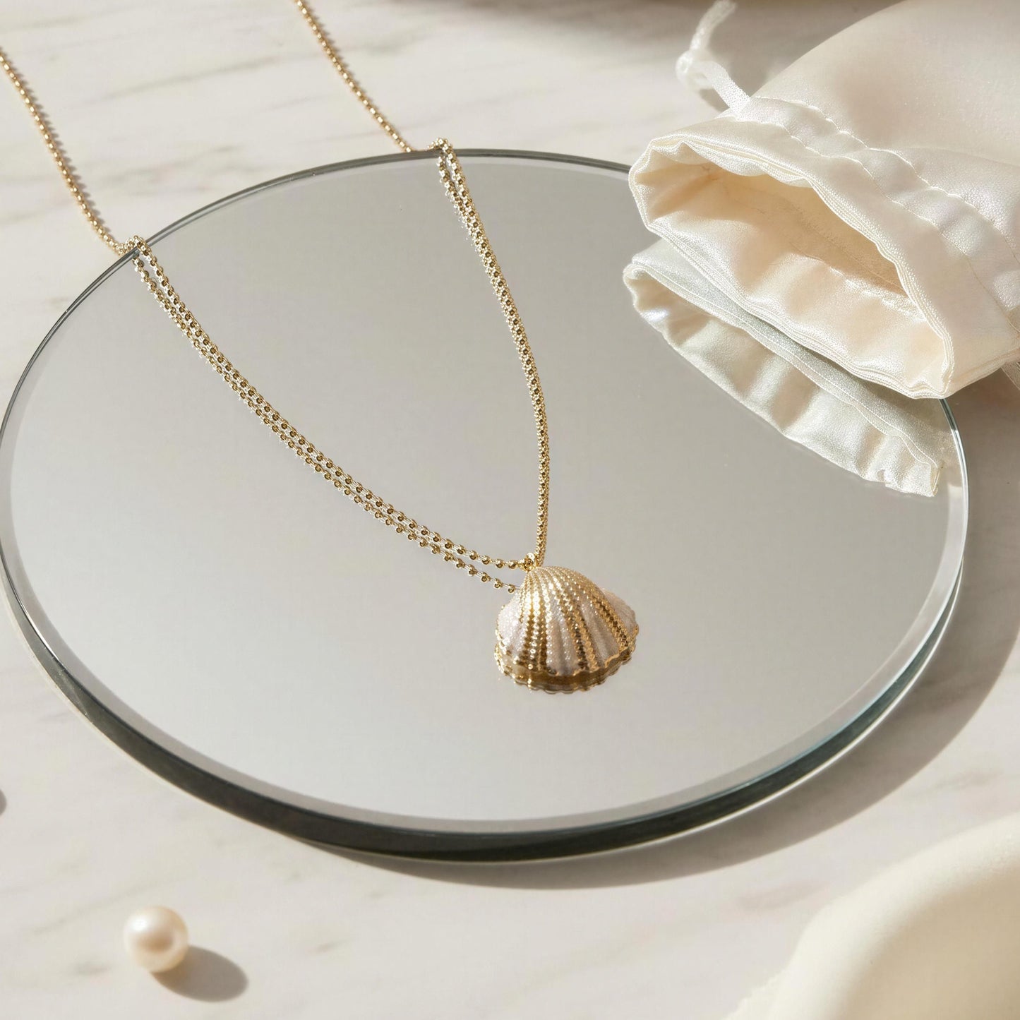 Gold shell pendant necklace on a round mirror with pearls and a white drawstring bag.