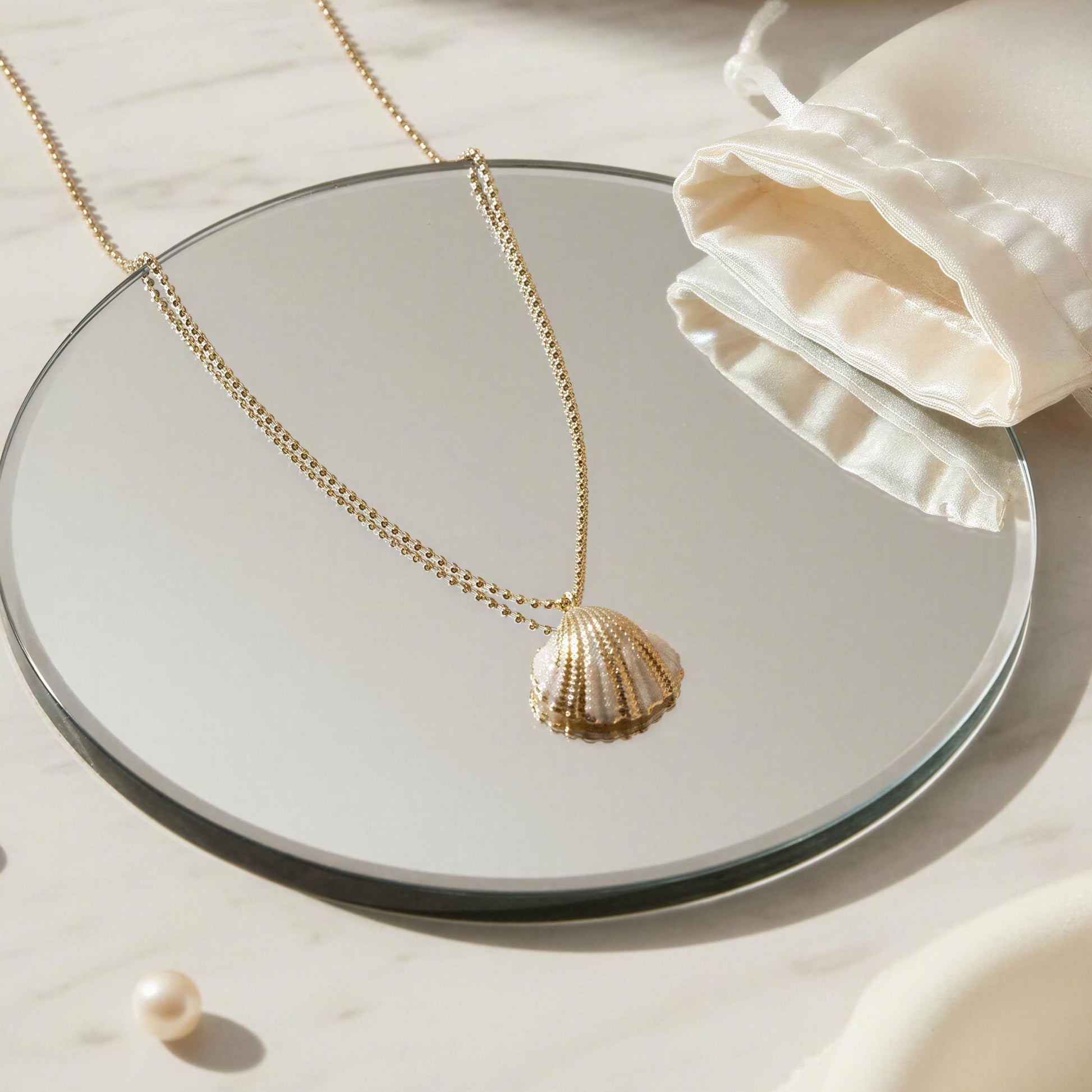 Gold shell pendant necklace on a round mirror with pearls and a white drawstring bag.