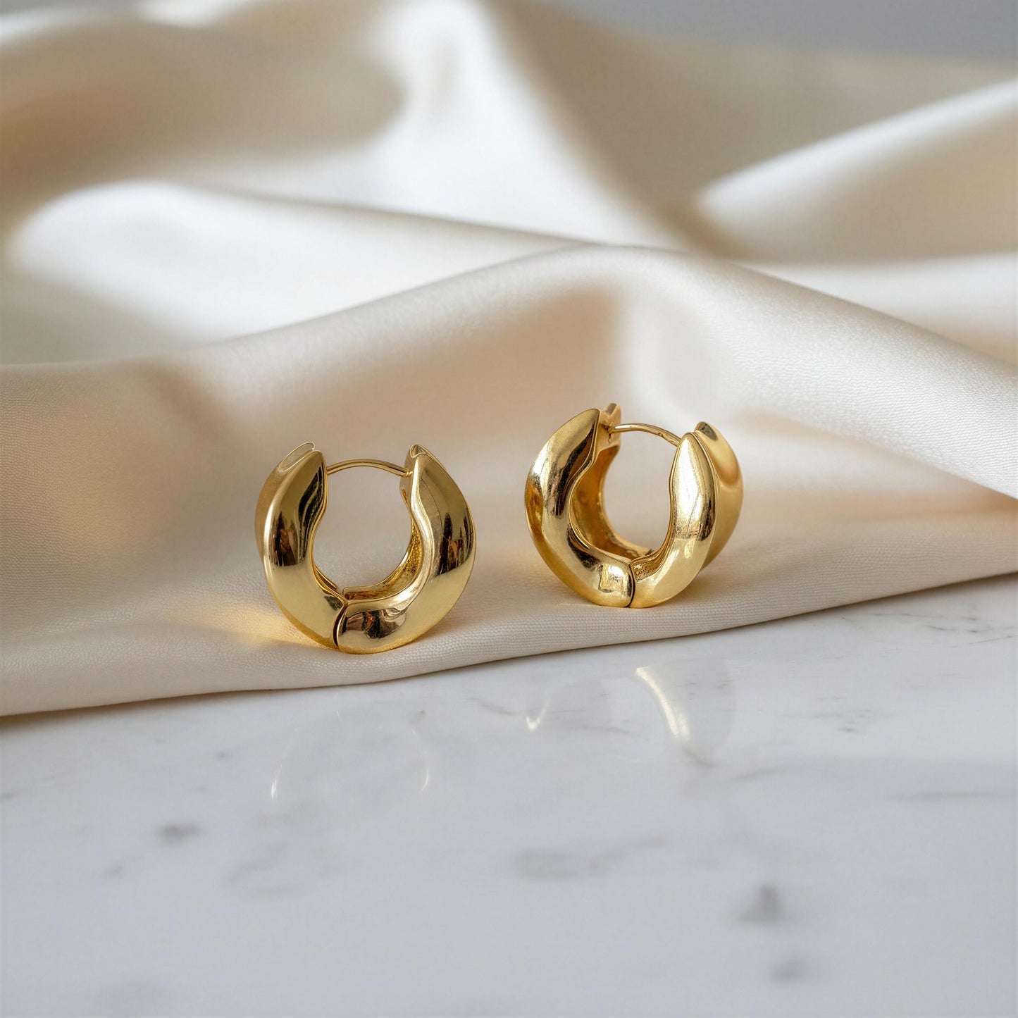The Isadora Hoops, a pair of chunky, polished gold huggie hoop earrings resting on draped cream satin fabric over a white marble surface.
