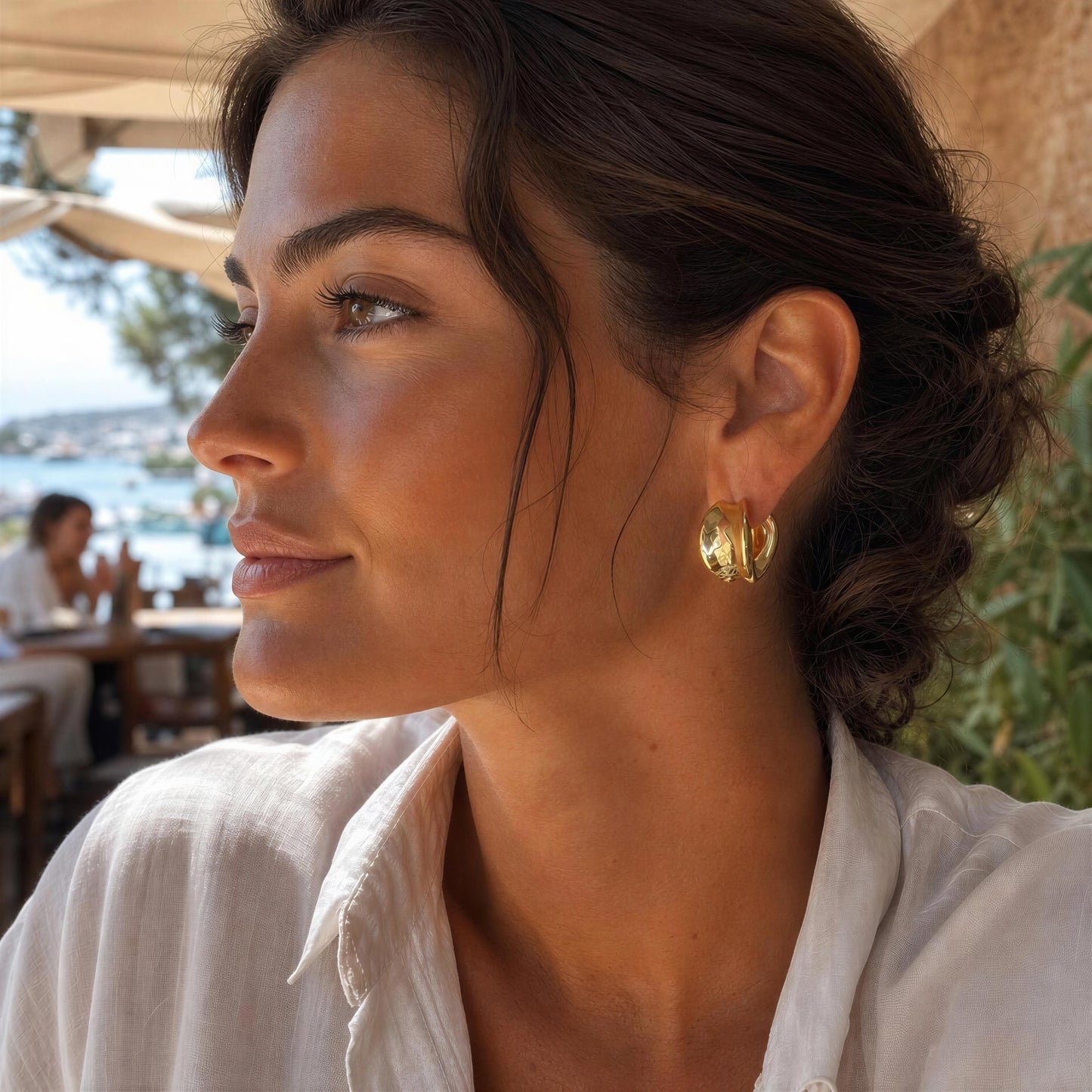 A close-up side profile of a woman wearing a pair of chunky gold hoop earrings at an outdoor cafe.