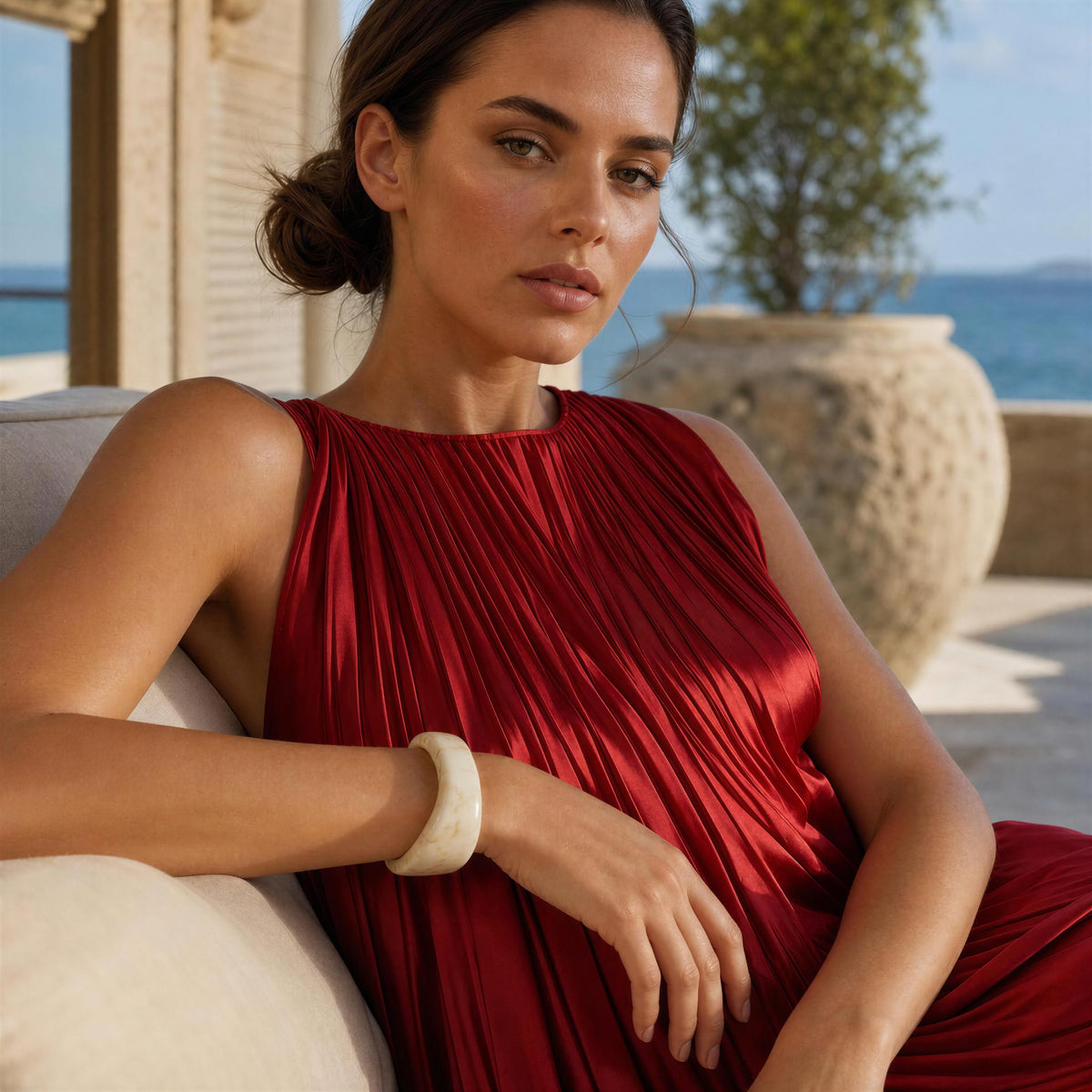 A woman wearing a sleeveless, pleated red dress sits on a light-colored outdoor sofa with a scenic background of a stone wall, a large planter, and the sea.