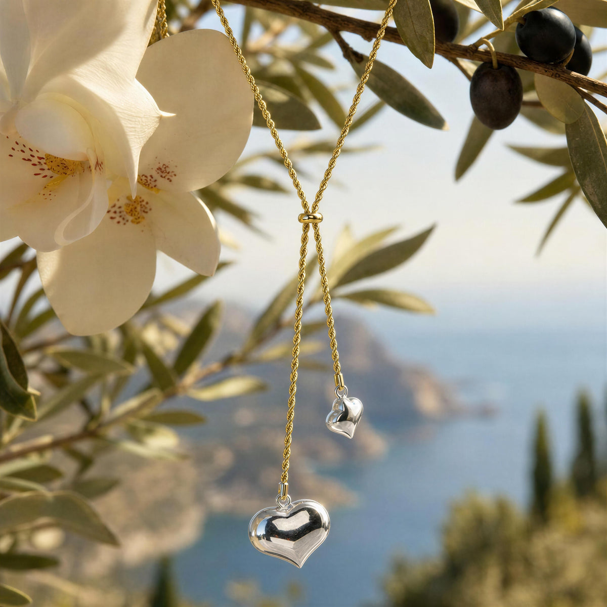 A gold-tone double-heart pendant necklace with a large polished silver heart and a smaller silver heart charm, hanging from olive branches with a coastal sea background.