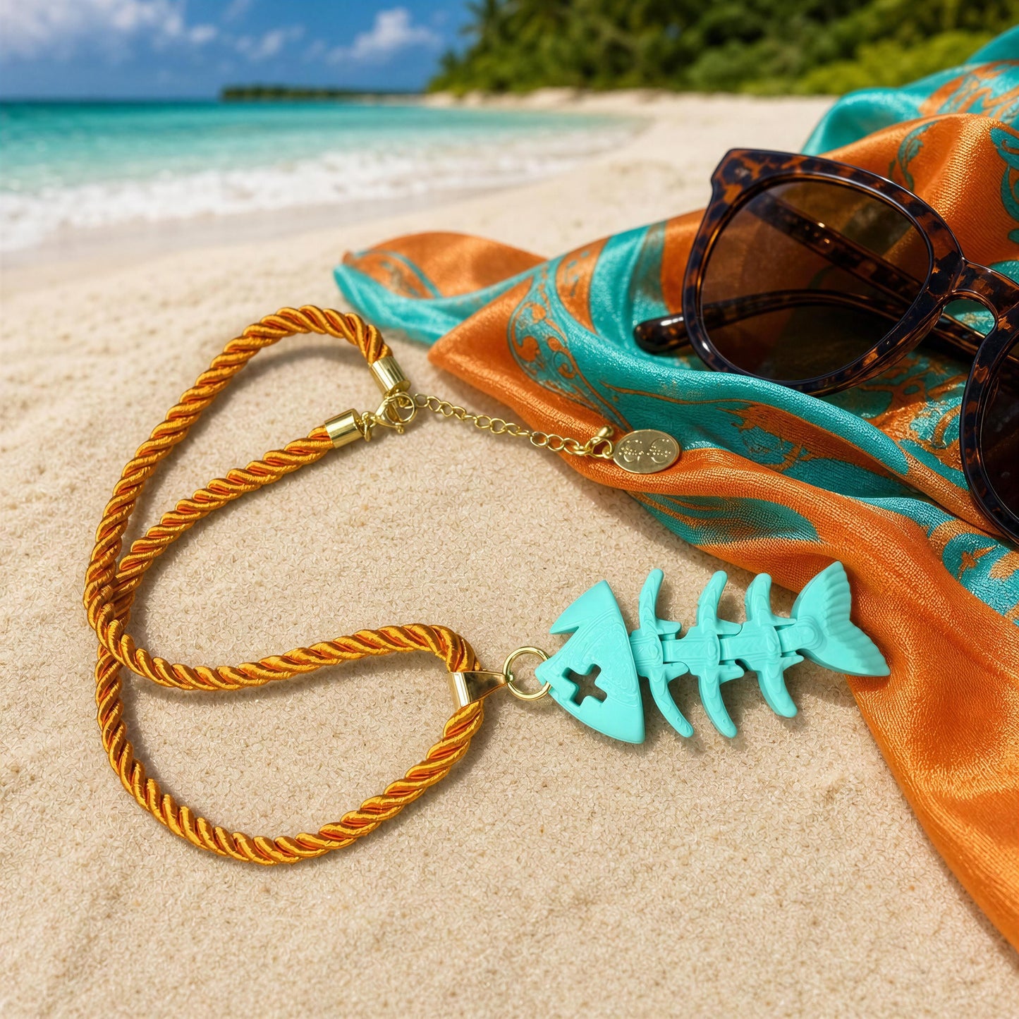 The Lisca Fishbone Necklace- Chica Chica Jewelry laid flat at Caribbean beach next to an orange and turquoise head scarf and tortoise sunglasses.