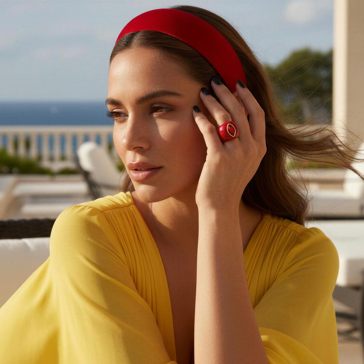 A woman wearing a bright red wide headband and a yellow top, posed outdoors with the ocean and lounge chairs blurred in the background. She is touching her face with one hand, which has dark nail polish and the The Lola Ring — Tomato-from Chica Chica Jewelry- Retro Glam Collection 
