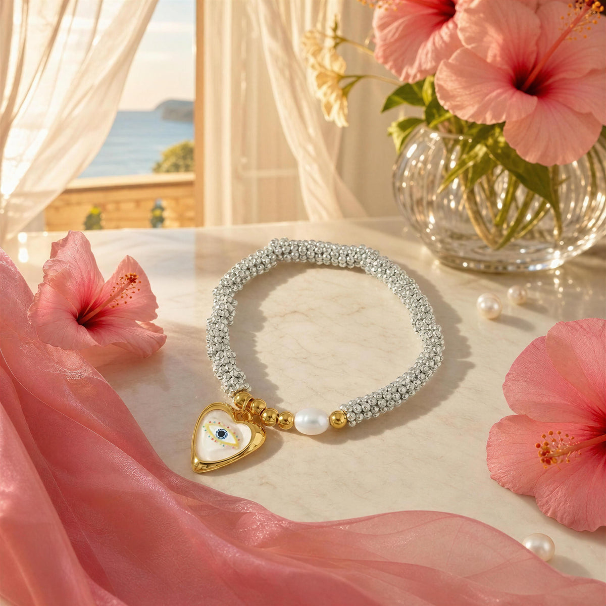 A beaded bracelet with a gold heart charm featuring an evil eye design, displayed on a marble surface with pink hibiscus flowers and a vase in the background.