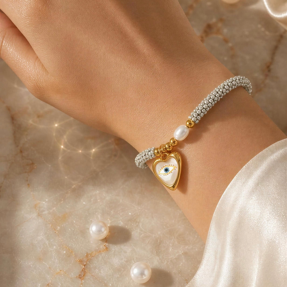 A close-up of a person's wrist wearing a delicate bracelet with a gold heart charm featuring an evil eye motif, set against a warm marble surface with scattered pearls.
