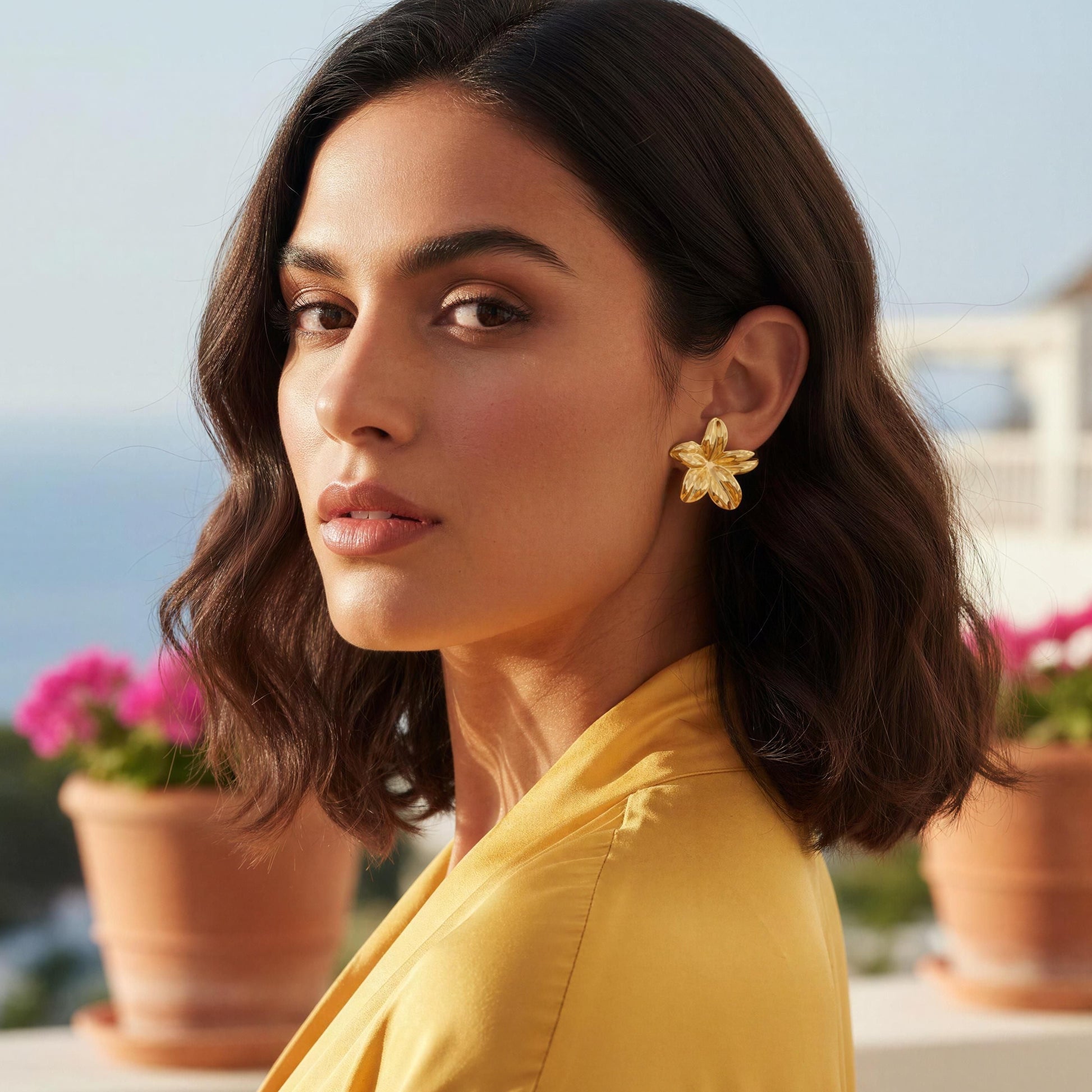 Woman wearing a yellow top with floral earrings, standing outdoors with plants and a blurred background