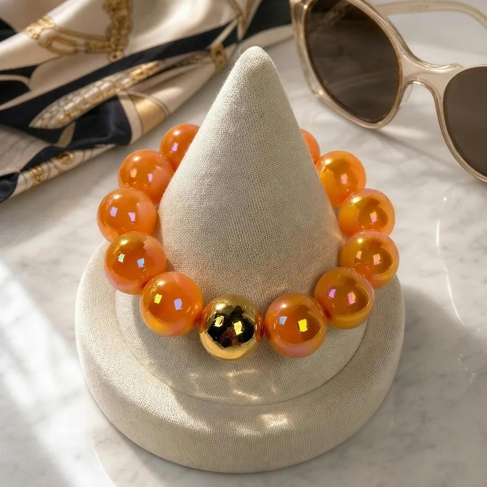 Orange beaded bracelet with a gold accent on a white stand, with sunglasses in the background.