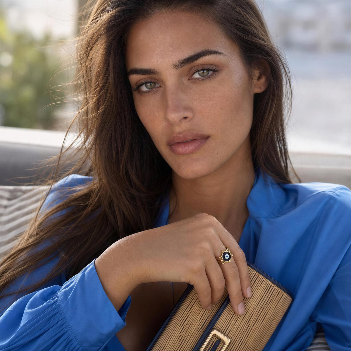 A close-up lifestyle shot of a woman with long brown hair wearing a bright blue blouse and holding a woven clutch, featuring a gold ring with a dark blue stone on her finger.