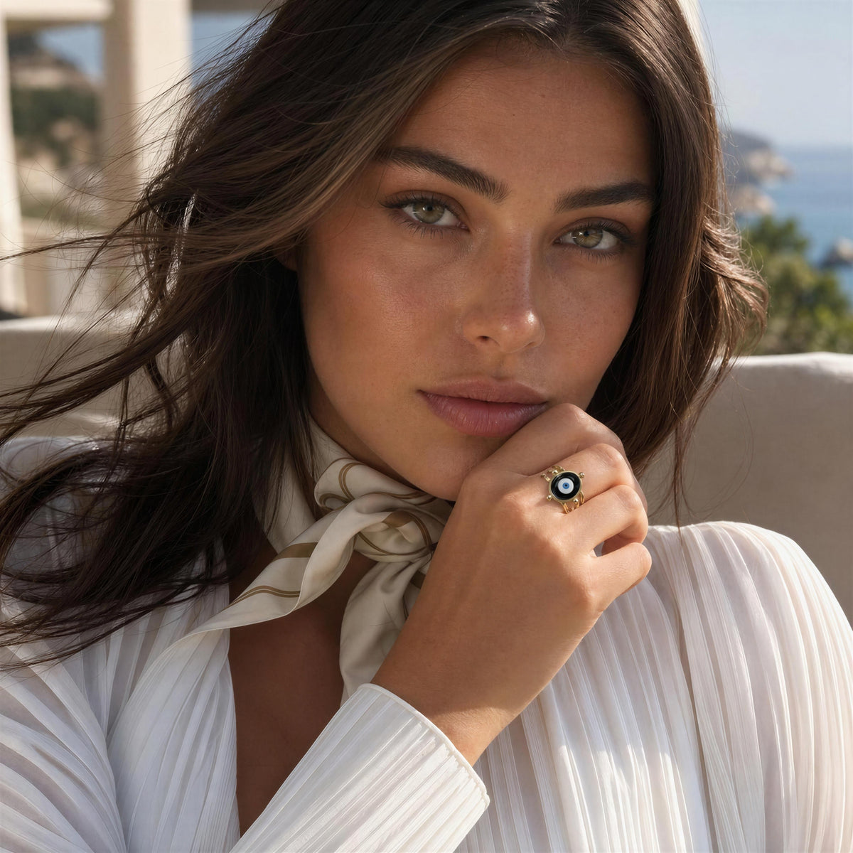 A close-up lifestyle shot of a woman wearing a white pleated top and a cream satin neck scarf, featuring a gold ring with a black and white evil eye design on her finger.