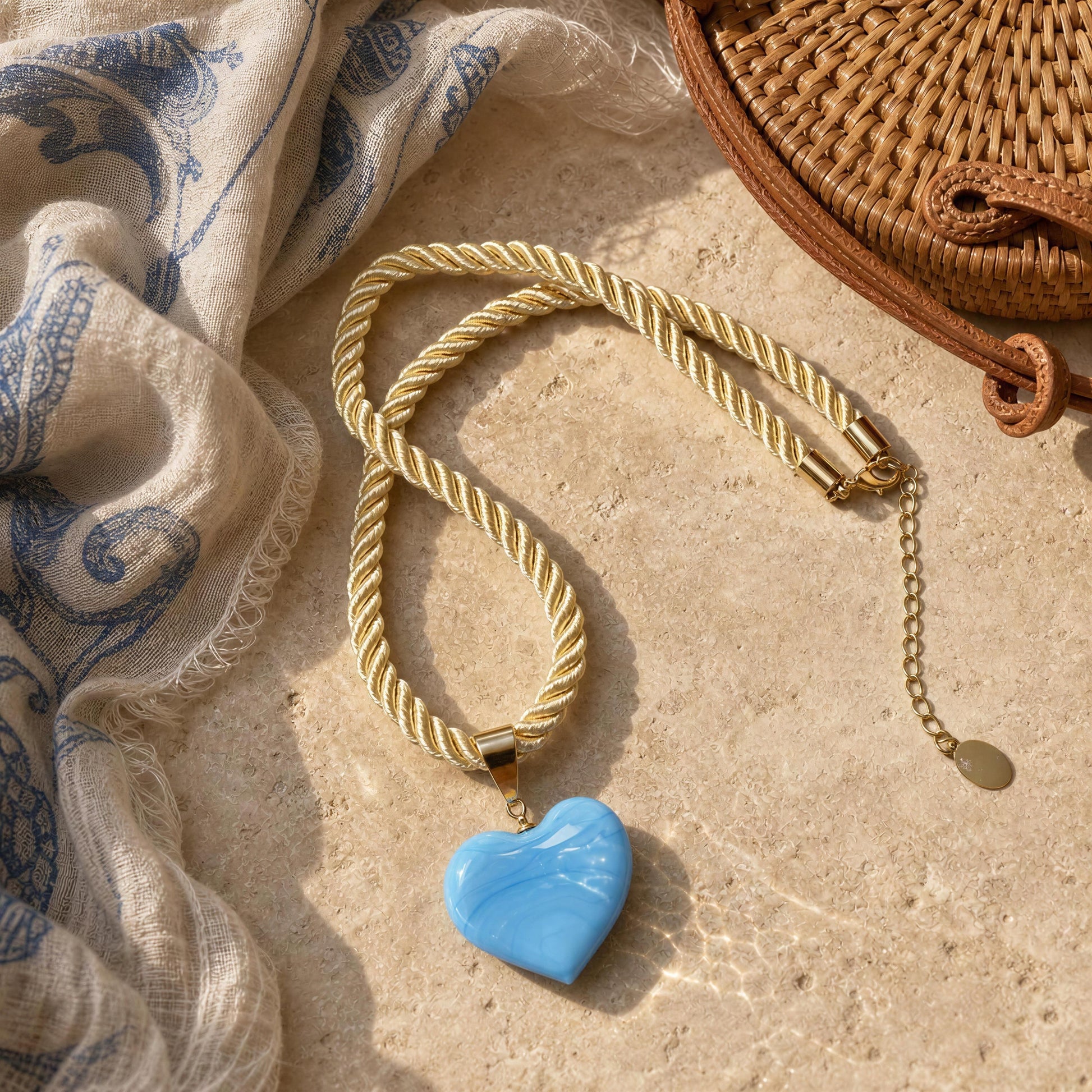 TheMavi Murano-style blue glass heart necklace with a soft braided cord and 18k gold-plated details by Chica Chica Jewelry next to rattan bag and scarf.