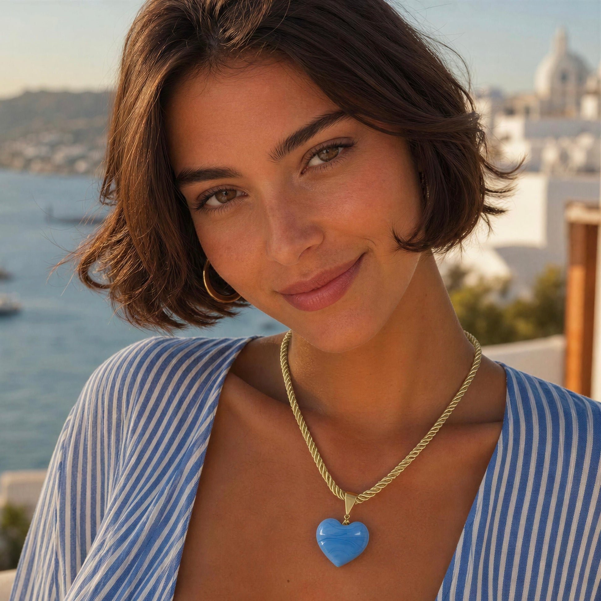 Model wearing the Mavi Murano-style blue glass heart necklace with a soft braided cord and 18k gold-plated details by Chica Chica Jewelry at a seaside restaurant overlooking the Bodrum coastline, Turkey.
