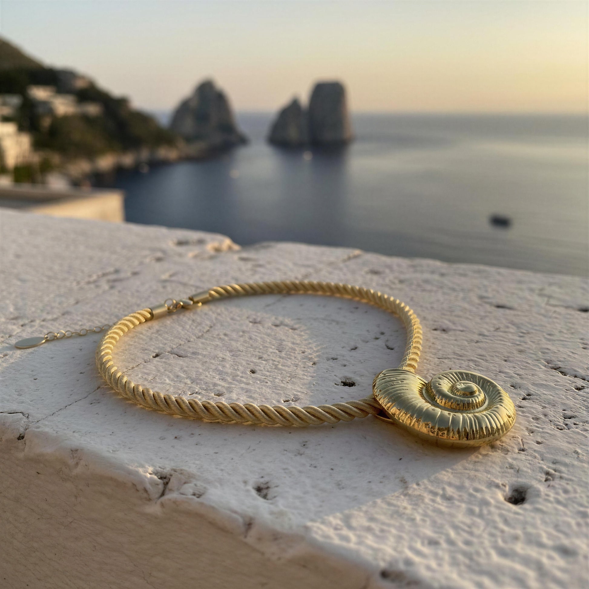 The Nautila Necklace from Chica Chica Jewelry,  a cream color rope necklace with a spiral shell-shaped pendant, resting on a white stone ledge with a blurred coastal background featuring the sea and rock formations.
