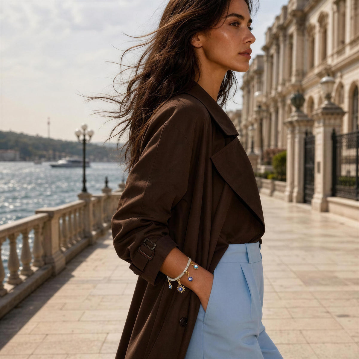 A woman in a dark brown trench coat and light blue trousers stands on a stone waterfront promenade with a classical building and water in the background wearing the Nazar Bracelet from Chica Chica Jewelry. 