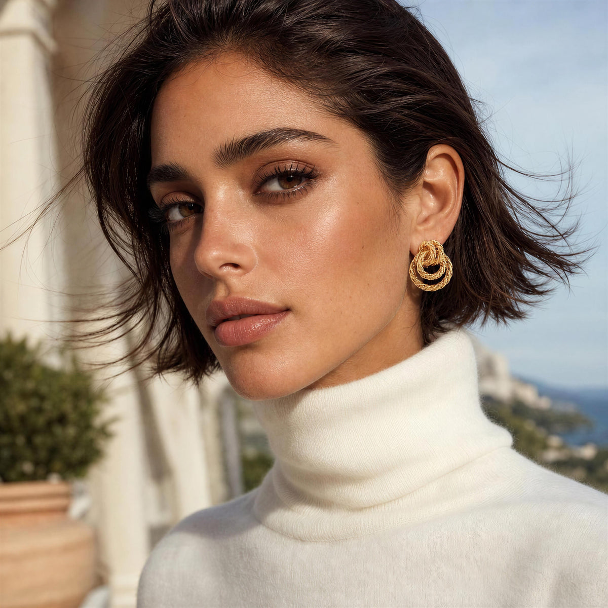 A close-up lifestyle shot of a woman with short dark hair wearing a white turtleneck sweater and the Nodo Earrings from Chica Chica Jewelry, a gold double-ring hoop earring, set against a sunny outdoor background with columns and a distant sea view.
