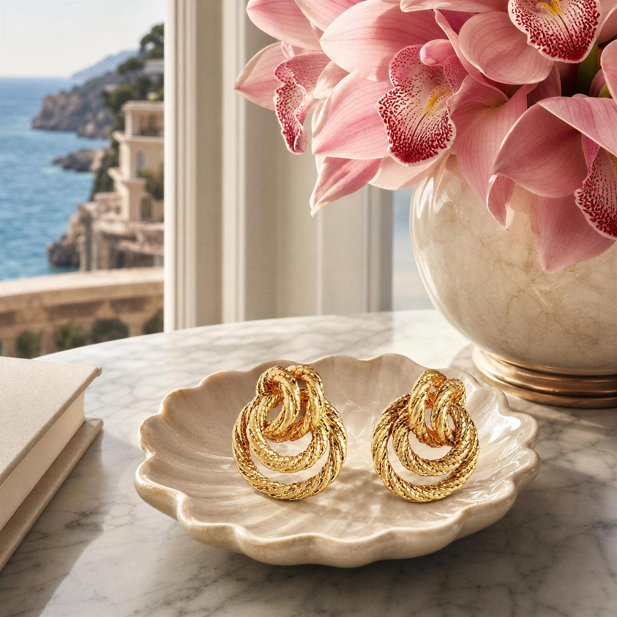 A pair of gold textured spiral hoop earrings displayed on a scalloped cream dish on a marble table, with a vase of pink orchids and a window view of a coastal town in the background.