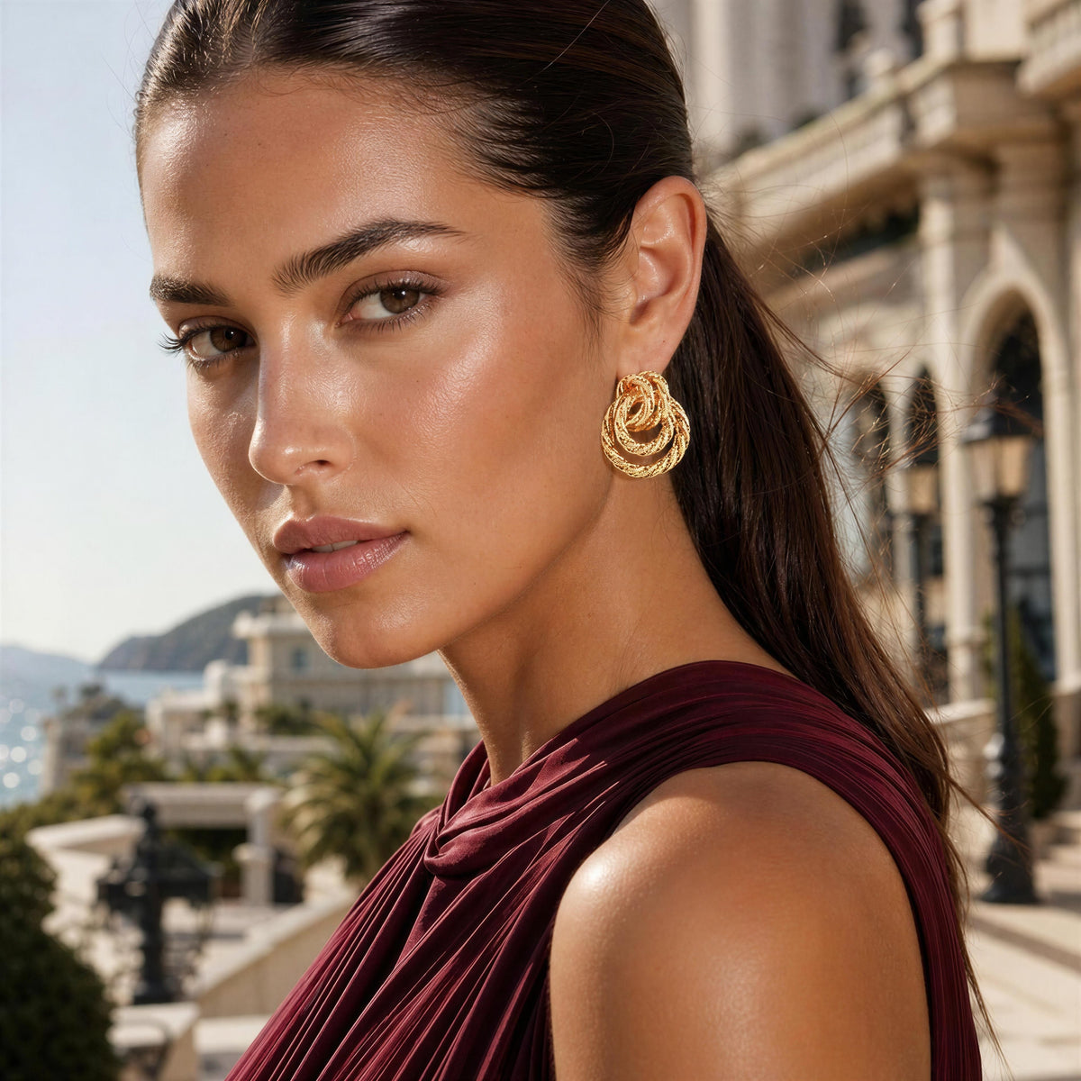 A woman in a burgundy dress wearing a gold spiral-shaped hoop earring, photographed outdoors with a blurred architectural background.