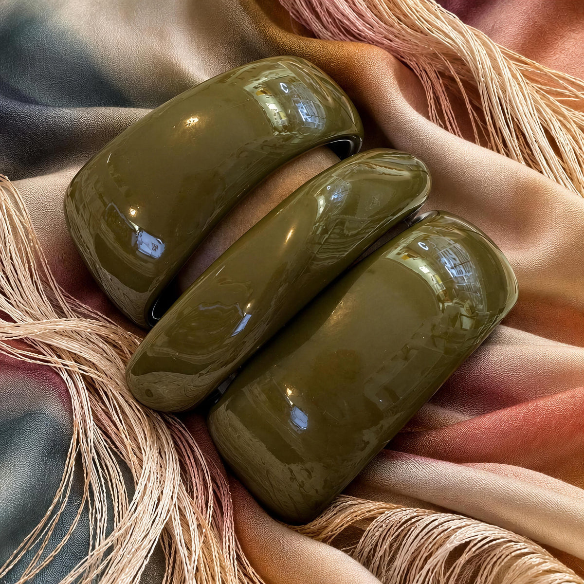 The Oliva Bangle Set from Chica Chica Jewelry. Three glossy olive green bangles arranged on draped satin fabric with beige tassels.