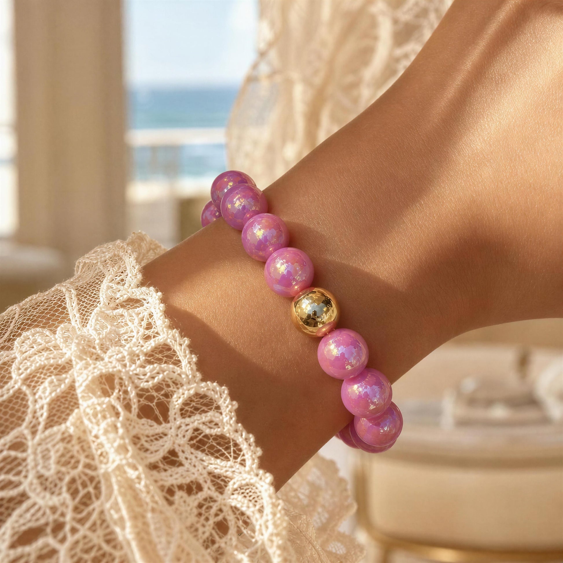 Pink beaded bracelet with a gold accent on a wrist, blurred background of a room with ocean view