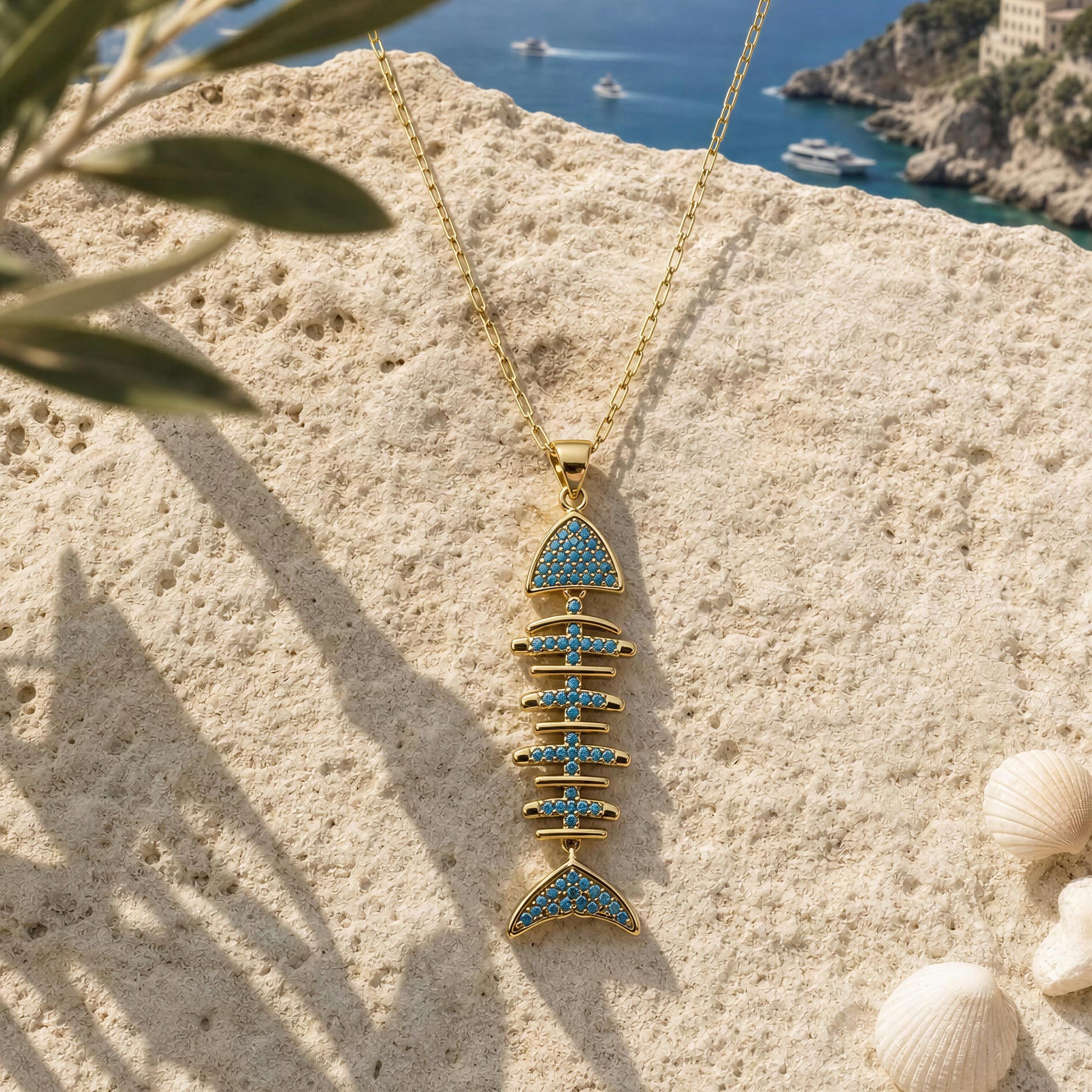 A gold fish skeleton pendant necklace with light blue stone accents, displayed on a textured beige stone surface with a coastal background showing the sea and cliffs.