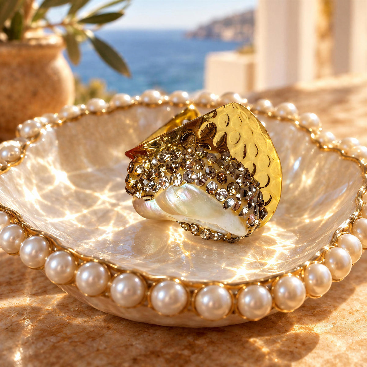 A gold and crystal-encrusted ring displayed on a pearl-bordered dish, with a sunlit beach and sea in the blurred background.