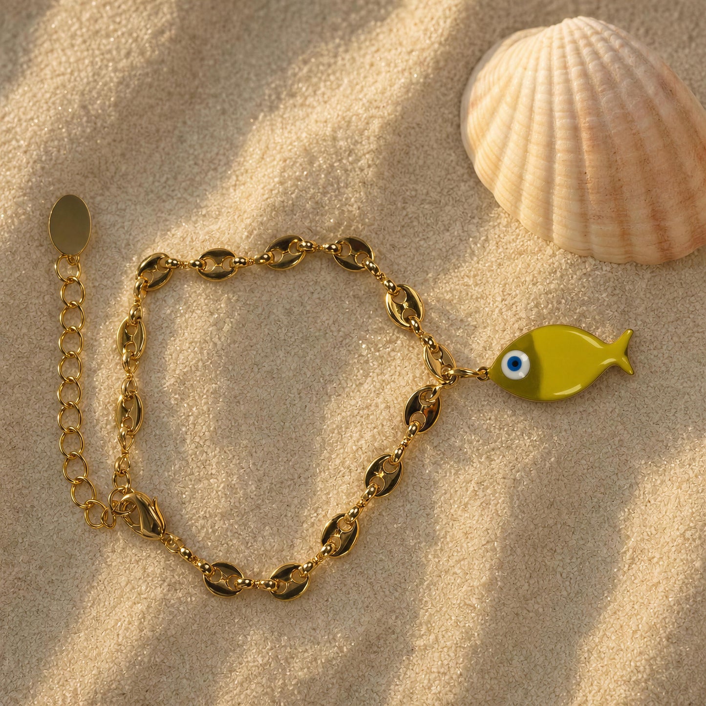 Gold bracelet with a fish charm on sand next to a shell