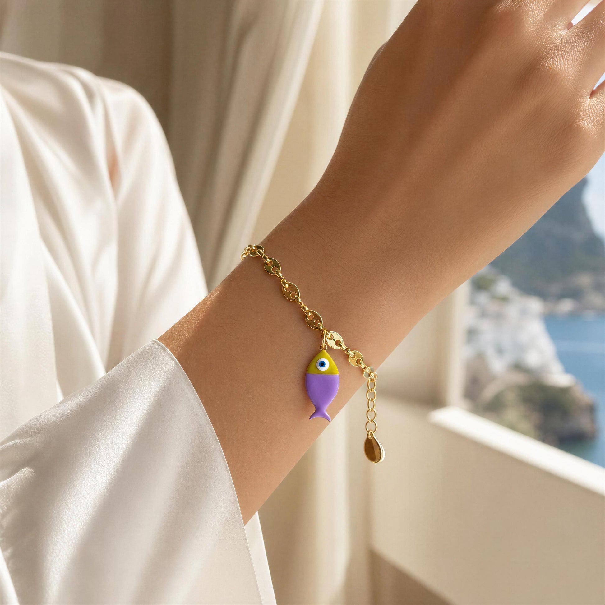 Gold bracelet with a purple fish charm on a wrist, with a blurred scenic background.