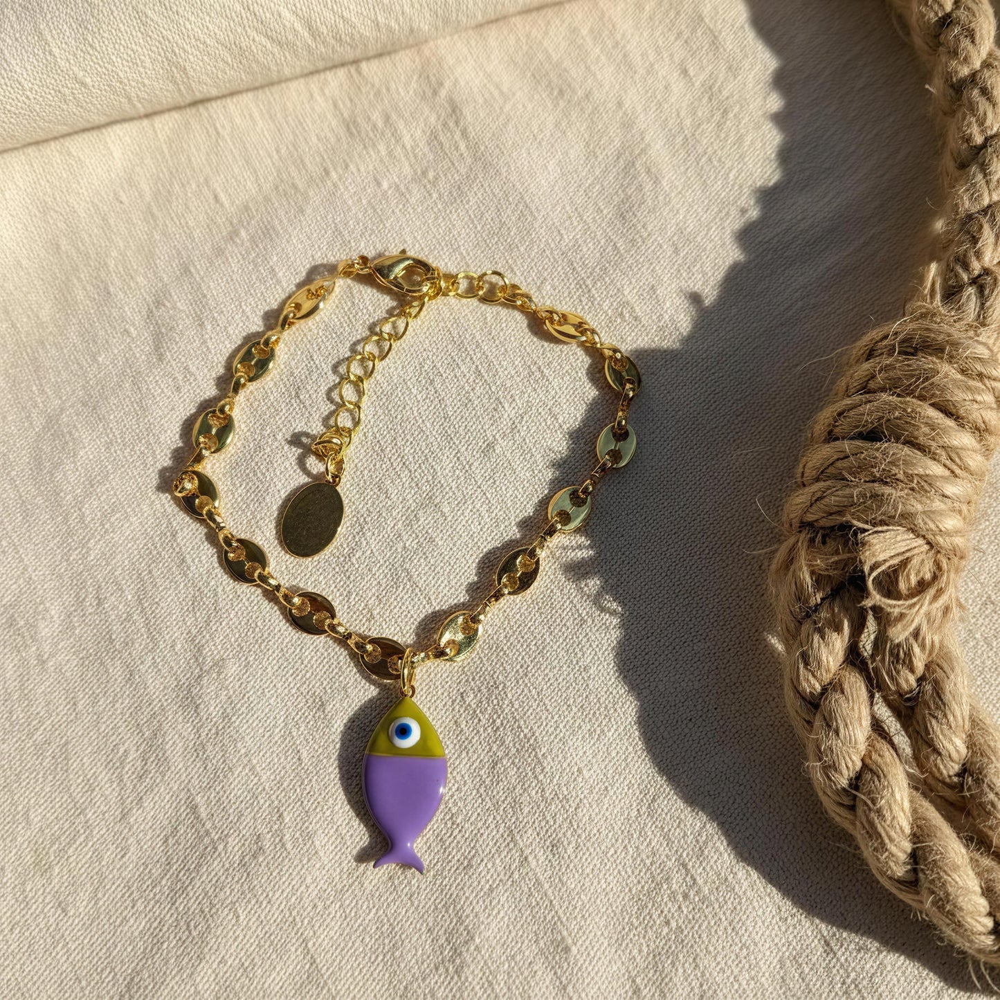 Gold bracelet with a purple fish charm on a textured beige surface