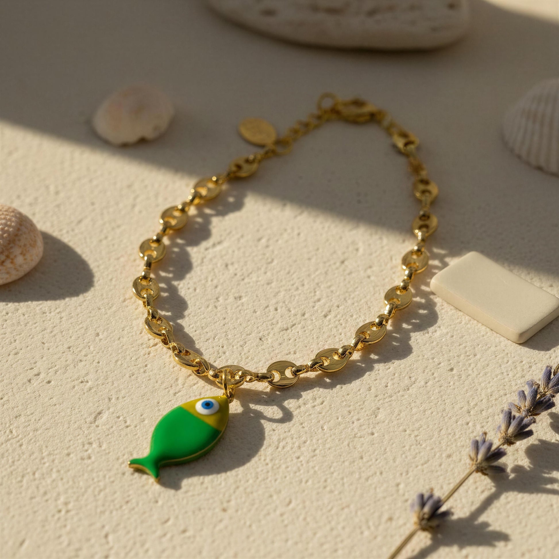 Gold bracelet with a green fish charm on a sandy surface with seashells and lavender.