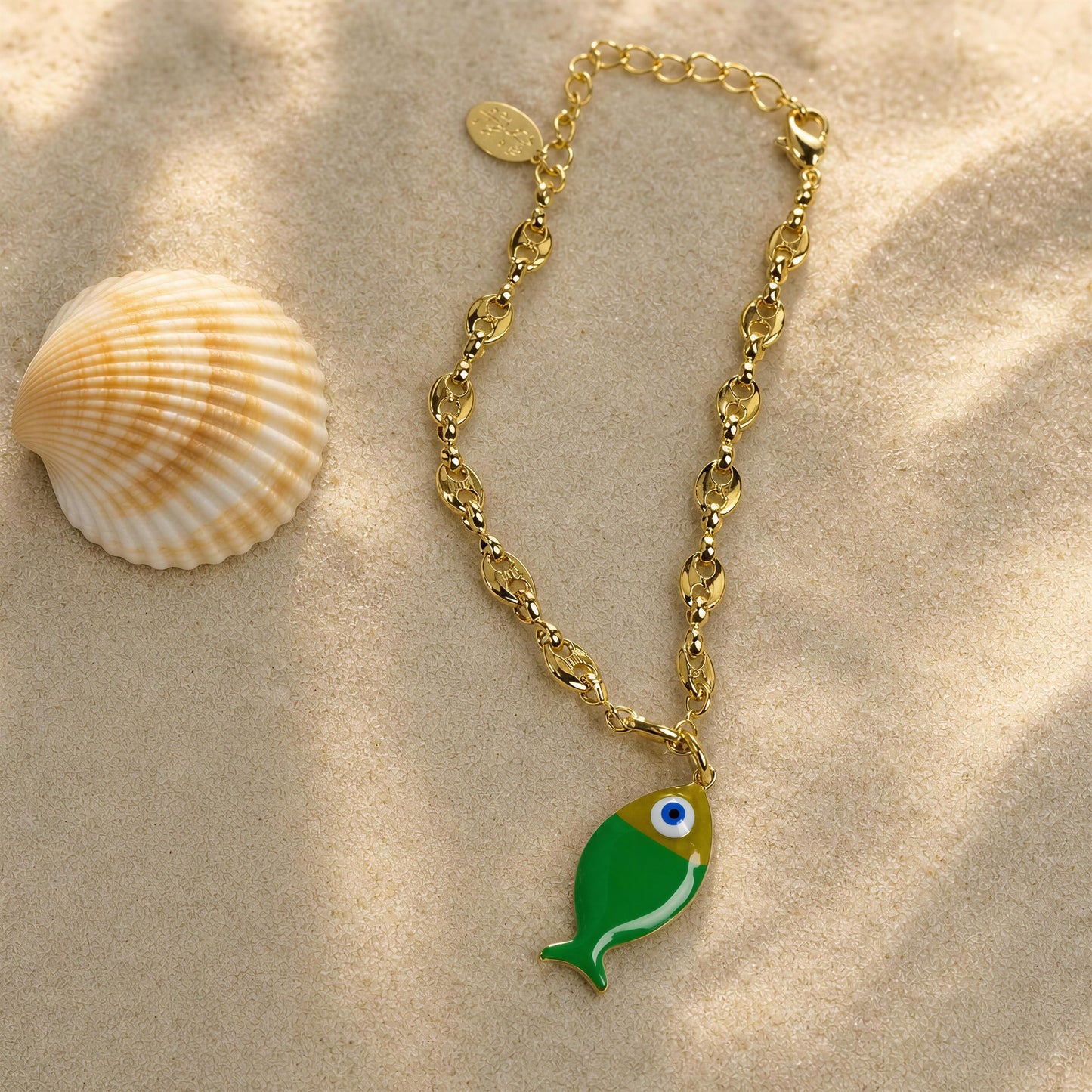 Gold bracelet with a green fish charm on sand with a shell