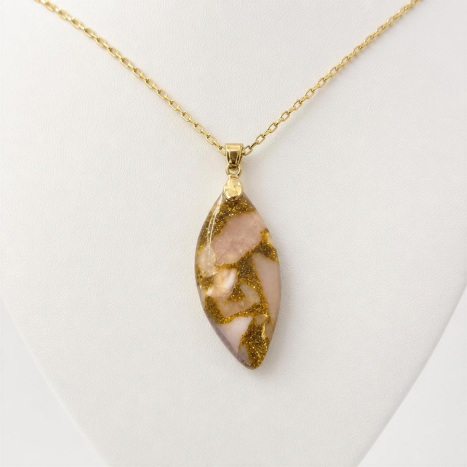A gold chain necklace with a marquise-shaped pendant featuring a pink stone inlay with gold glitter, displayed on a white jewelry bust.
