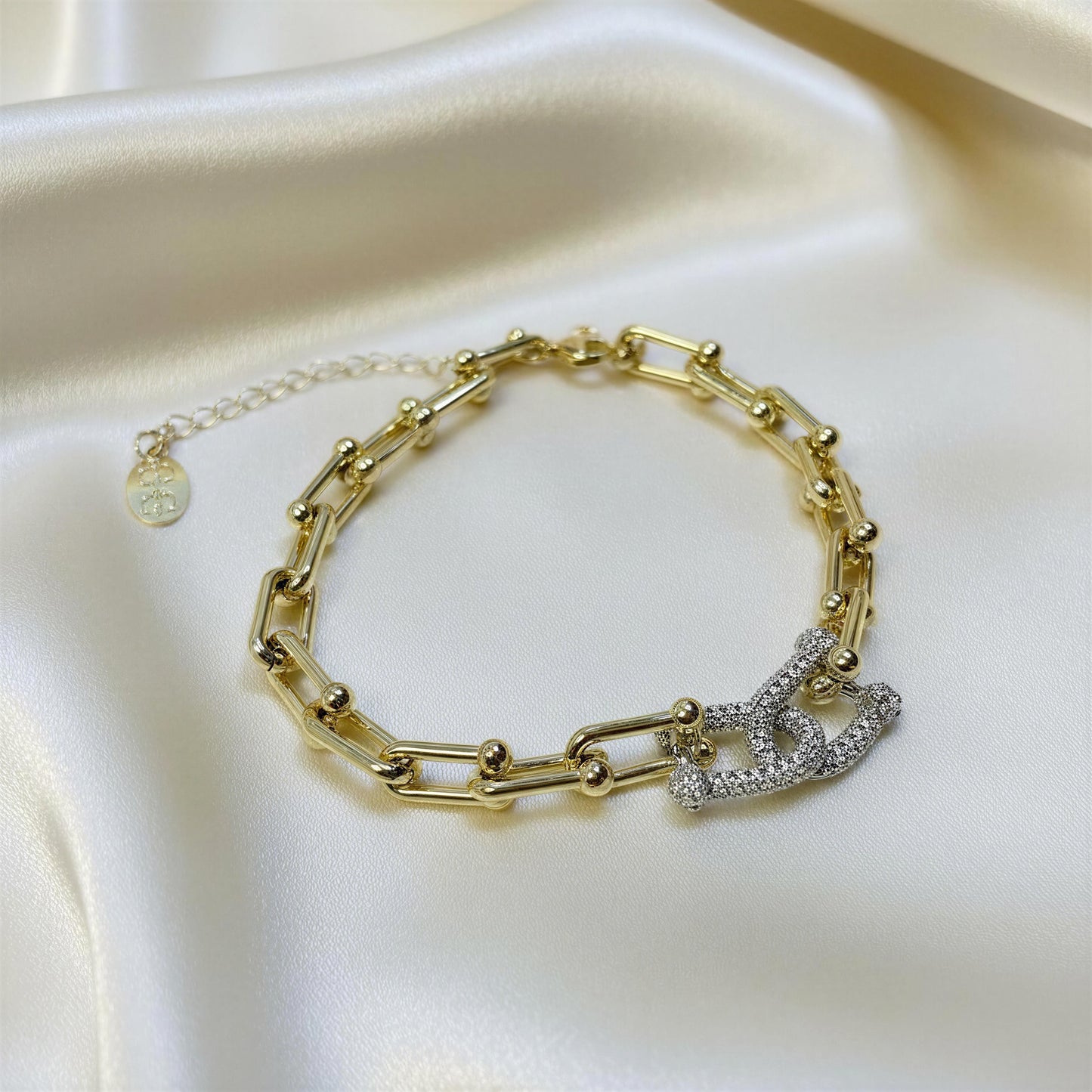 The Radiance Link Bracelet, a gold chain link bracelet with a polished interlocking double-C link charm paved with clear crystals, displayed on a draped cream satin fabric.