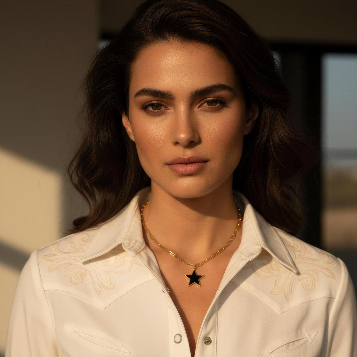 A close-up portrait of a woman wearing a white western-style shirt with embroidered details and a gold necklace featuring a black star pendant, set against a softly blurred indoor background.