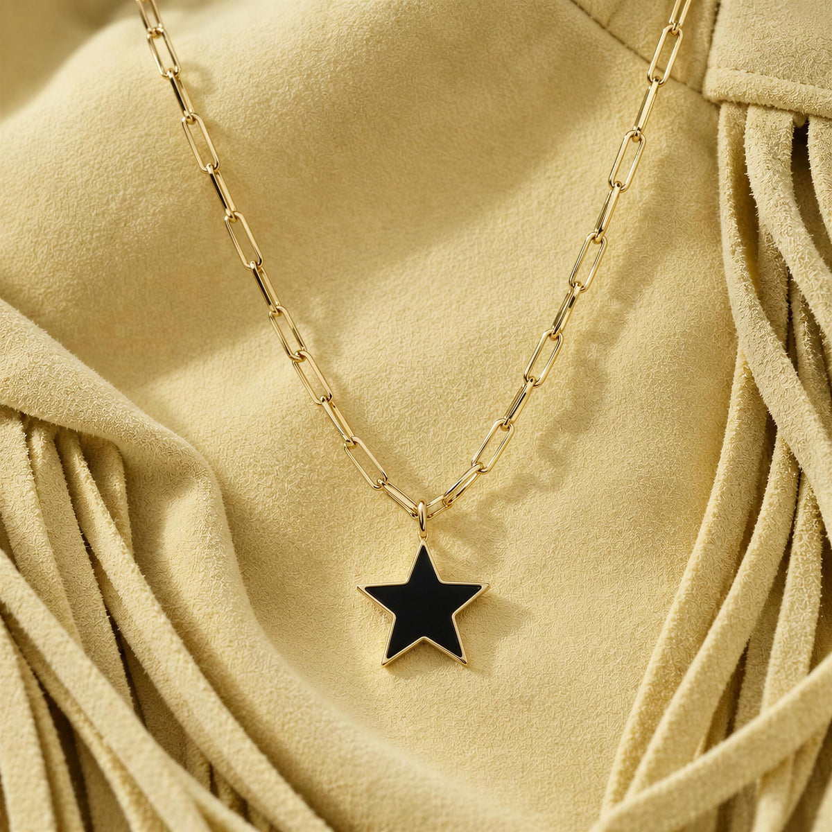 A gold paperclip chain necklace with a black star pendant, displayed on a draped beige suede fabric background.