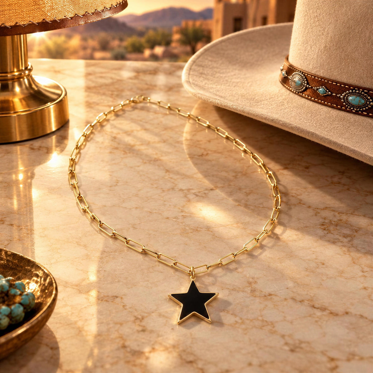 A gold chain necklace with a black star pendant is displayed on a warm-toned marble surface, styled with a cowboy hat, a lamp, and turquoise beads in a desert landscape setting.
