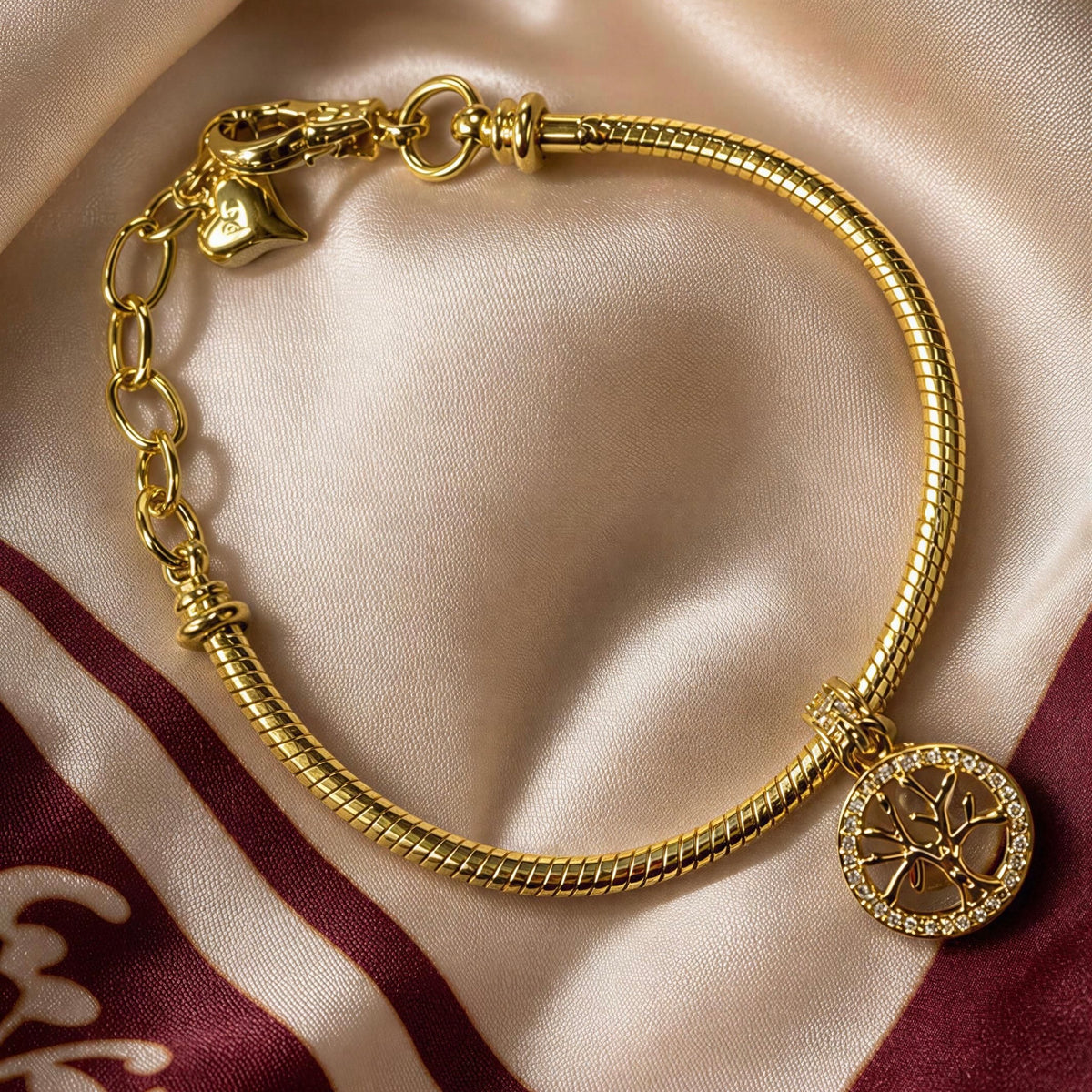 A gold snake chain bracelet with a tree of life pendant, displayed on a beige satin fabric with a maroon patterned corner.