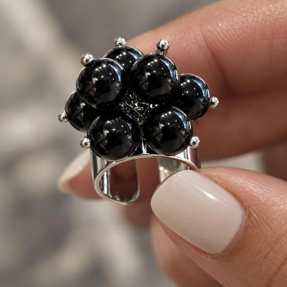 The Sabina Ring from Chica Chica Jewelry, a  silver-toned ring with a cluster of glossy black round stones, held between fingers against a softly blurred gray background.