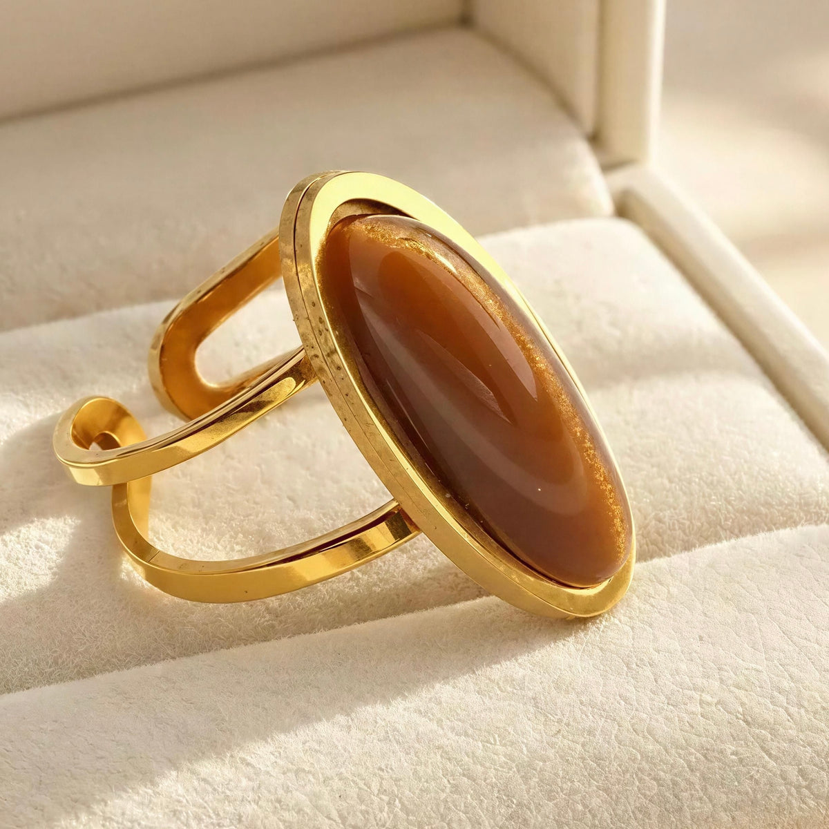 The Sahara Ring- Chica Chica Jewelry. A gold-toned open-band ring with a large polished oval brown stone, photographed resting on a cream-colored jewelry box cushion.