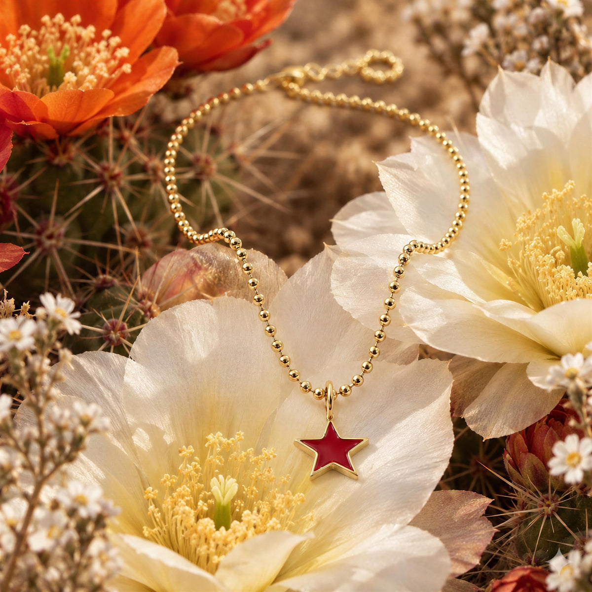 A gold beaded necklace with a red star pendant, styled on white cactus flowers with orange blossoms and cacti in the background.