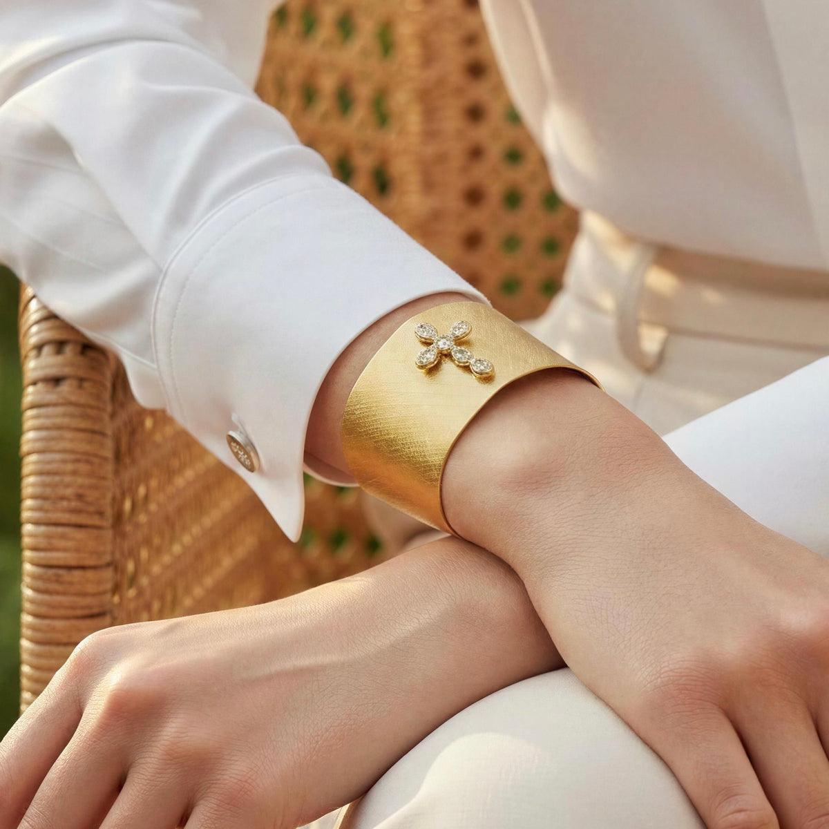 A close-up of a person wearing the Serafina Cuff from Chica Chica Jewelry,  a wide, polished gold cuff bracelet with a small floral crystal design on the front, paired with a white long-sleeve shirt.