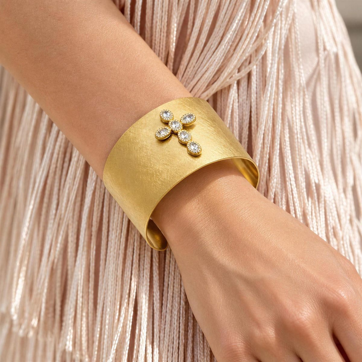 A close-up of a person's wrist wearing the Serafina Cuff from Chica Chica Jewelry, a wide textured gold cuff bracelet with a small diamond-encrusted floral motif at the front, against a pale pink fringed garment.