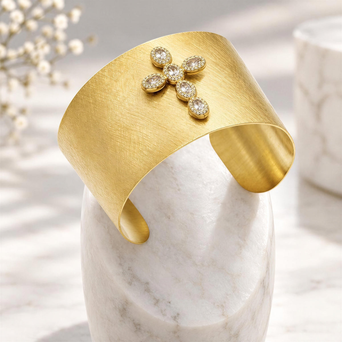 The Serafina Cuff- Chica Chica Jewelry. A wide brushed gold cuff bracelet with a flower-shaped cluster of small clear stones at the front, displayed on a white marble stand.