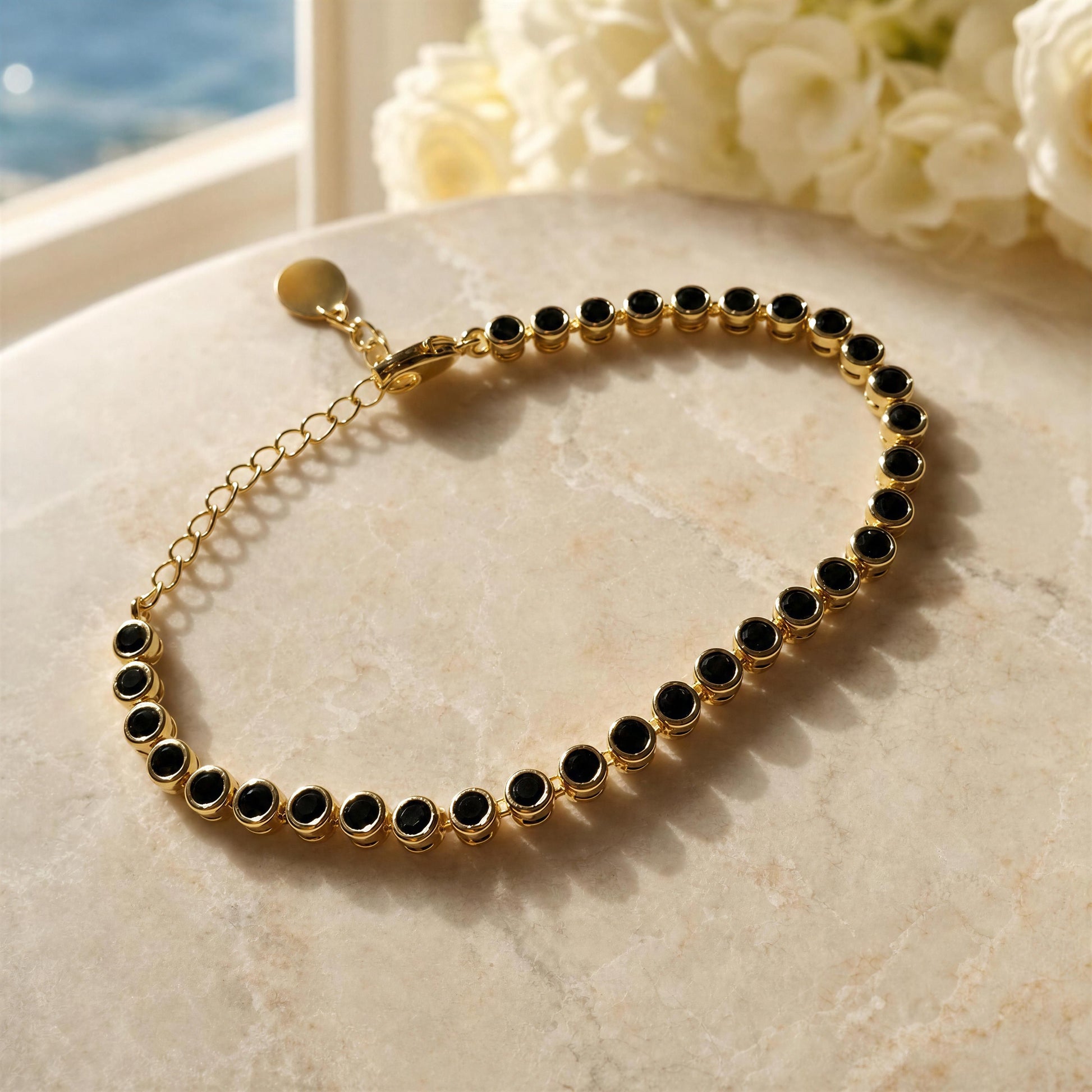 The Siena Black Tennis Bracelet displayed on a light beige stone surface with soft-focus white flowers and a window in the background.