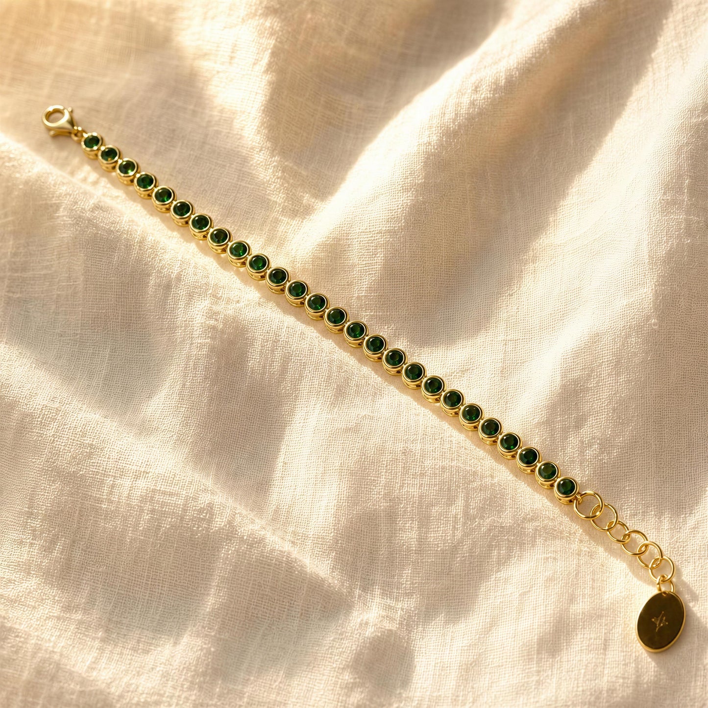 The Siena Emerald Tennis Bracelet, laid diagonally on a textured cream linen fabric background.