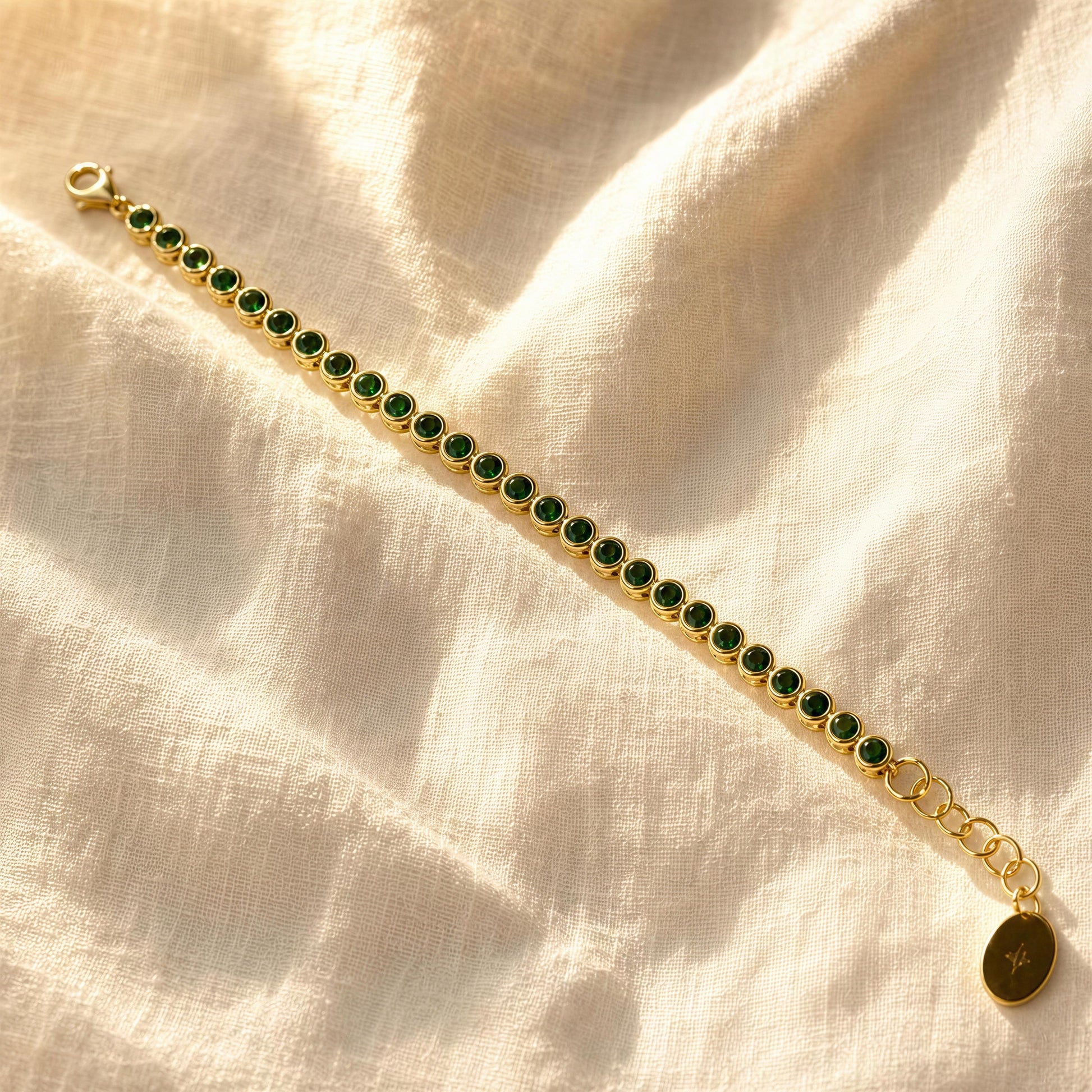 The Siena Emerald Tennis Bracelet, laid diagonally on a textured cream linen fabric background.
